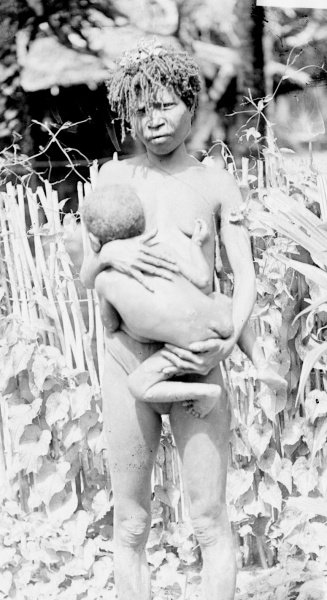 Fotografie. Stojící žena drží v náručí dítě (už dost velké), které kojí. Nová Guinea, 1896